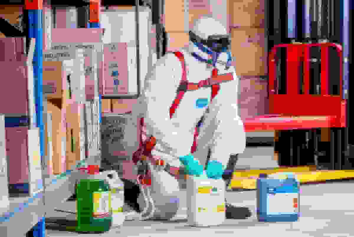 Worker in full protective hazmat suit and respirator handling chemical containers in warehouse, demonstrating proper workplace safety PPE protocols for hazardous material management